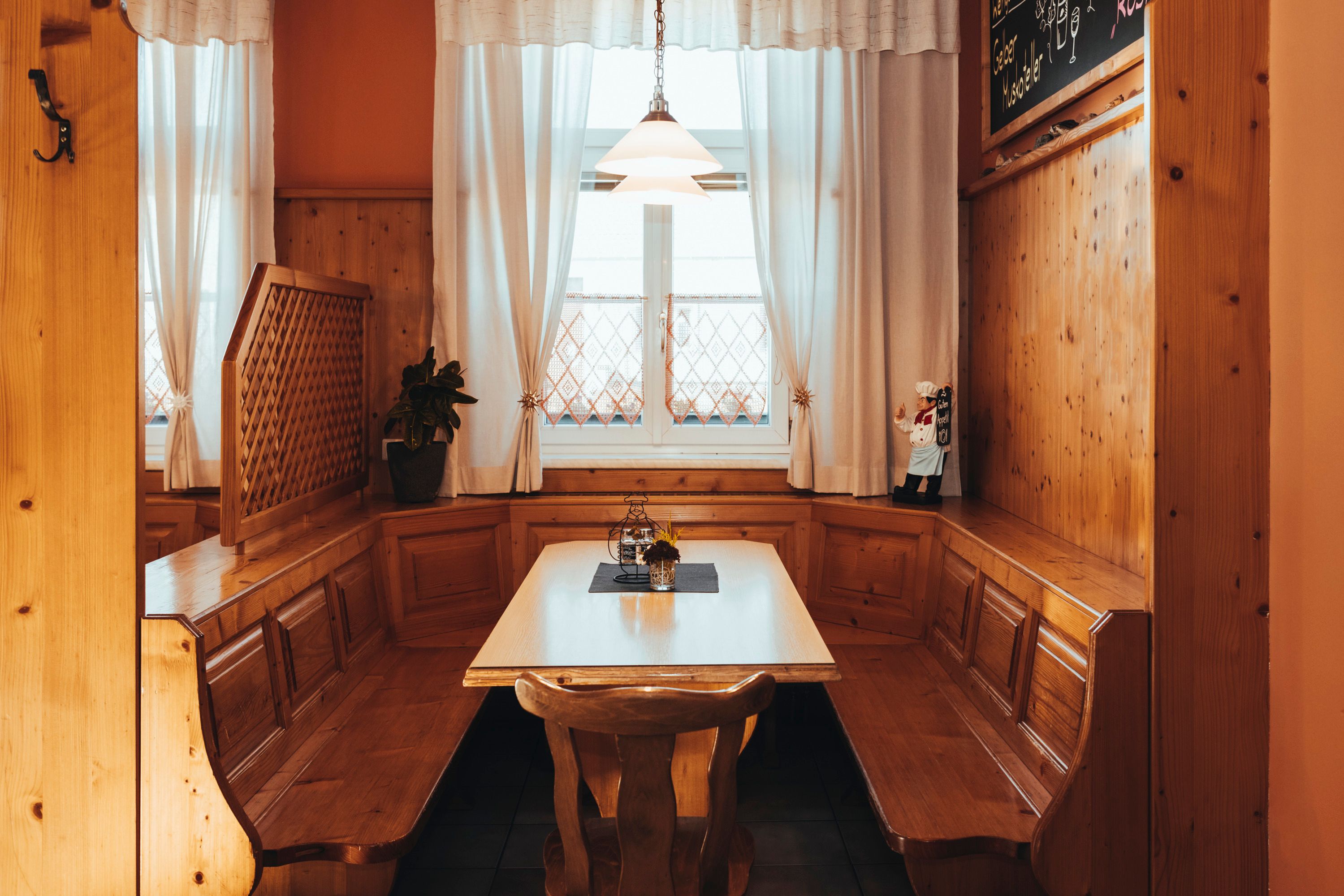 Gemütliche Holzeckbank mit Tisch in einem rustikalen Raum, dekoriert mit Vorhängen und einer kleinen Figur.