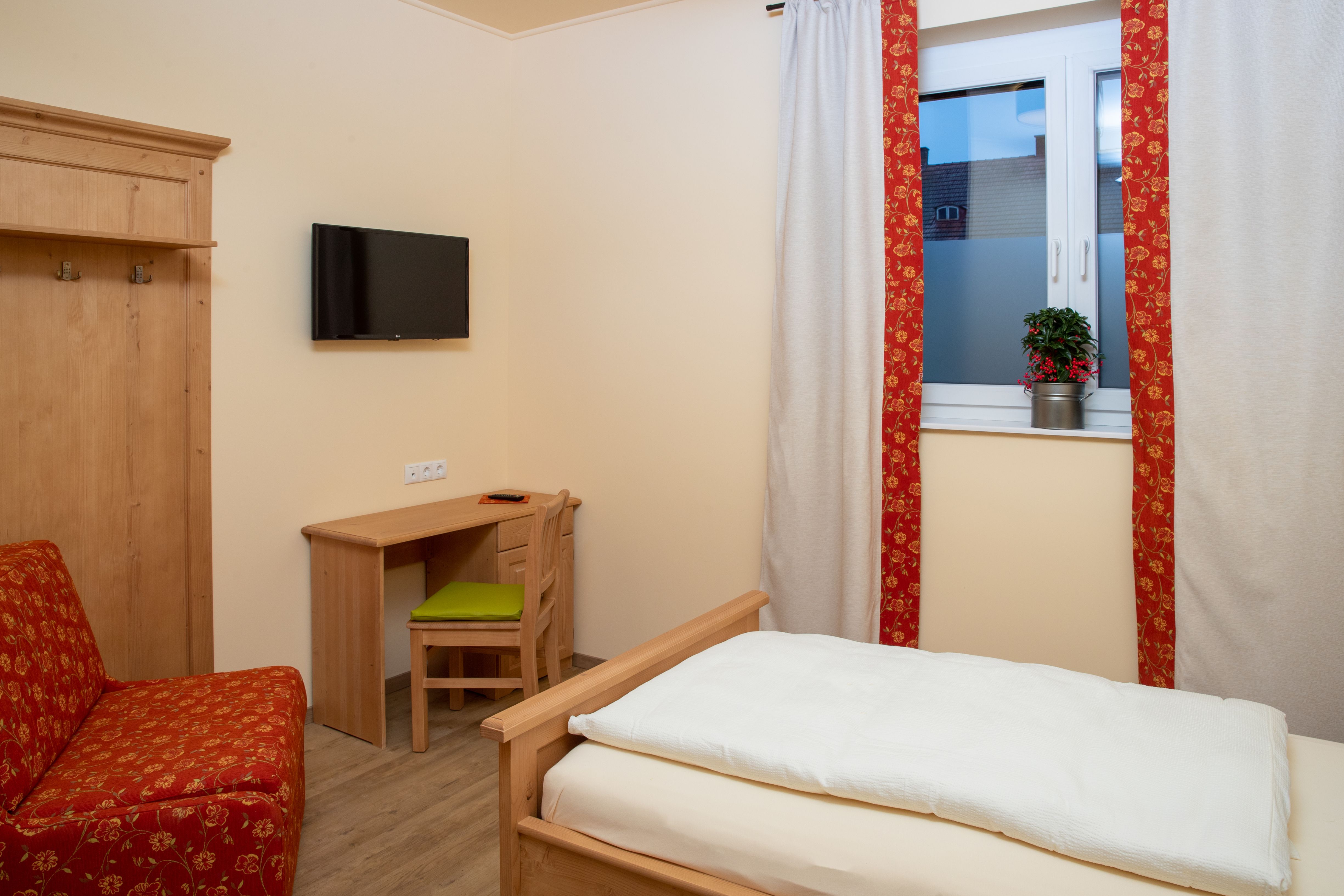 Ein gemütliches Einzelzimmer mit Holzmöbeln, einem Bett, einem Schreibtisch und einem Fernseher an der Wand. Ein Fenster mit Vorhängen und einer Pflanze.