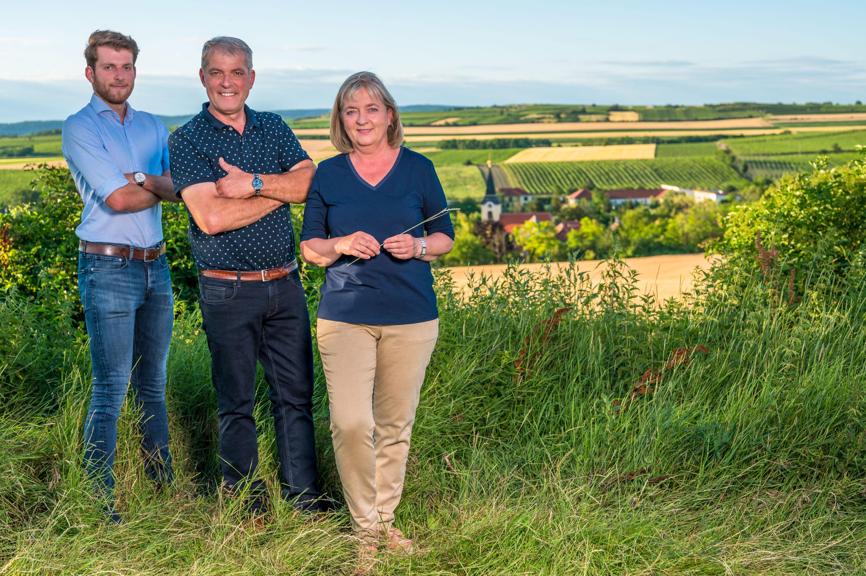 Drei Personen stehen in einem Weingarten mit Blick auf eine ländliche Landschaft im Hintergrund.