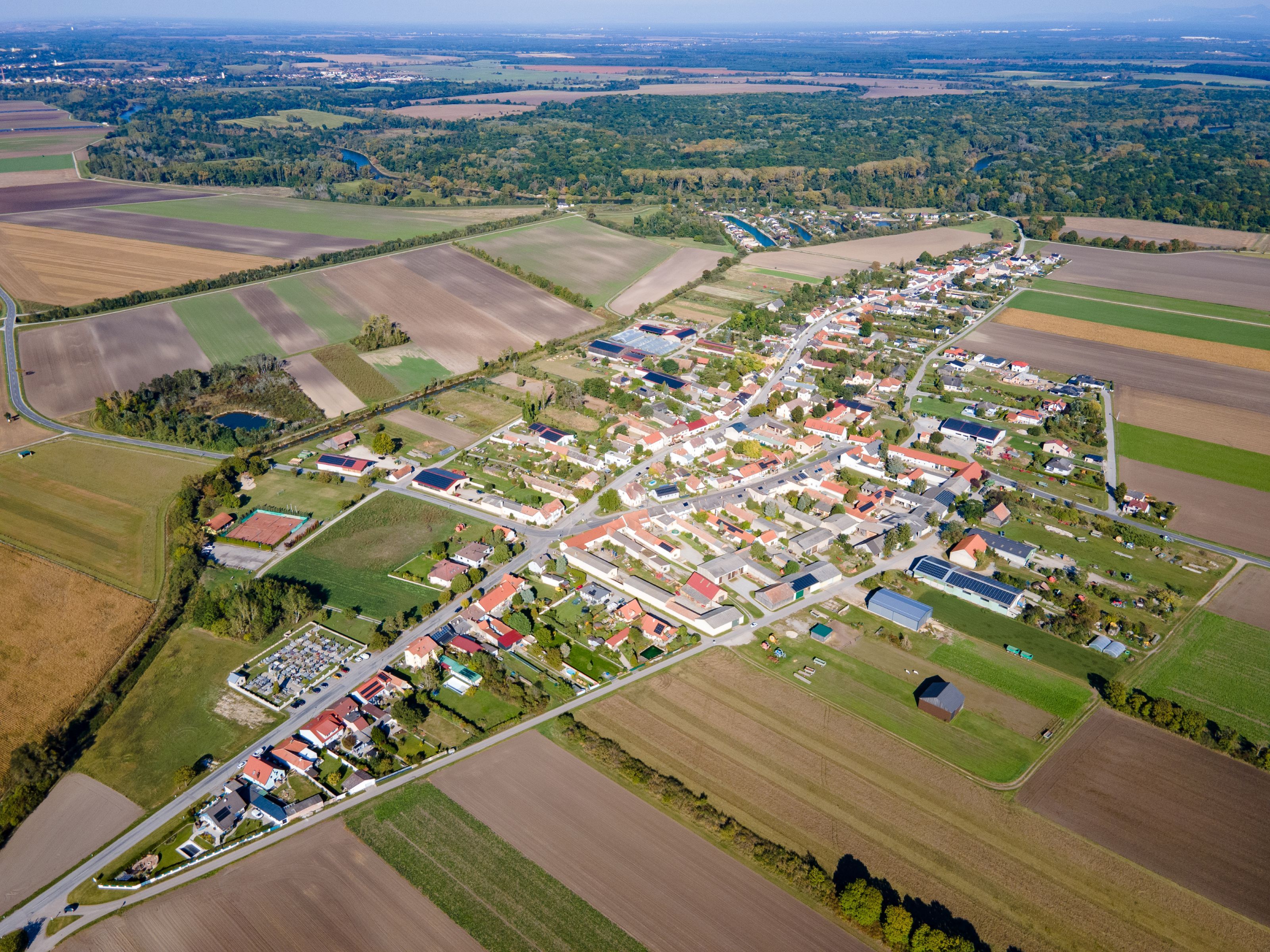 Luftaufnahme der Gemeinde Weiden an der March mit Feldern, Wohnhäusern und dem Flusslauf im Hintergrund.