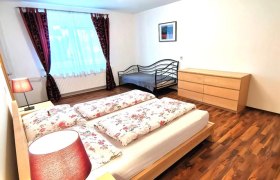 Schlafzimmer mit Doppelbett, Einzelbett, Kommode und Blumen in Vase.