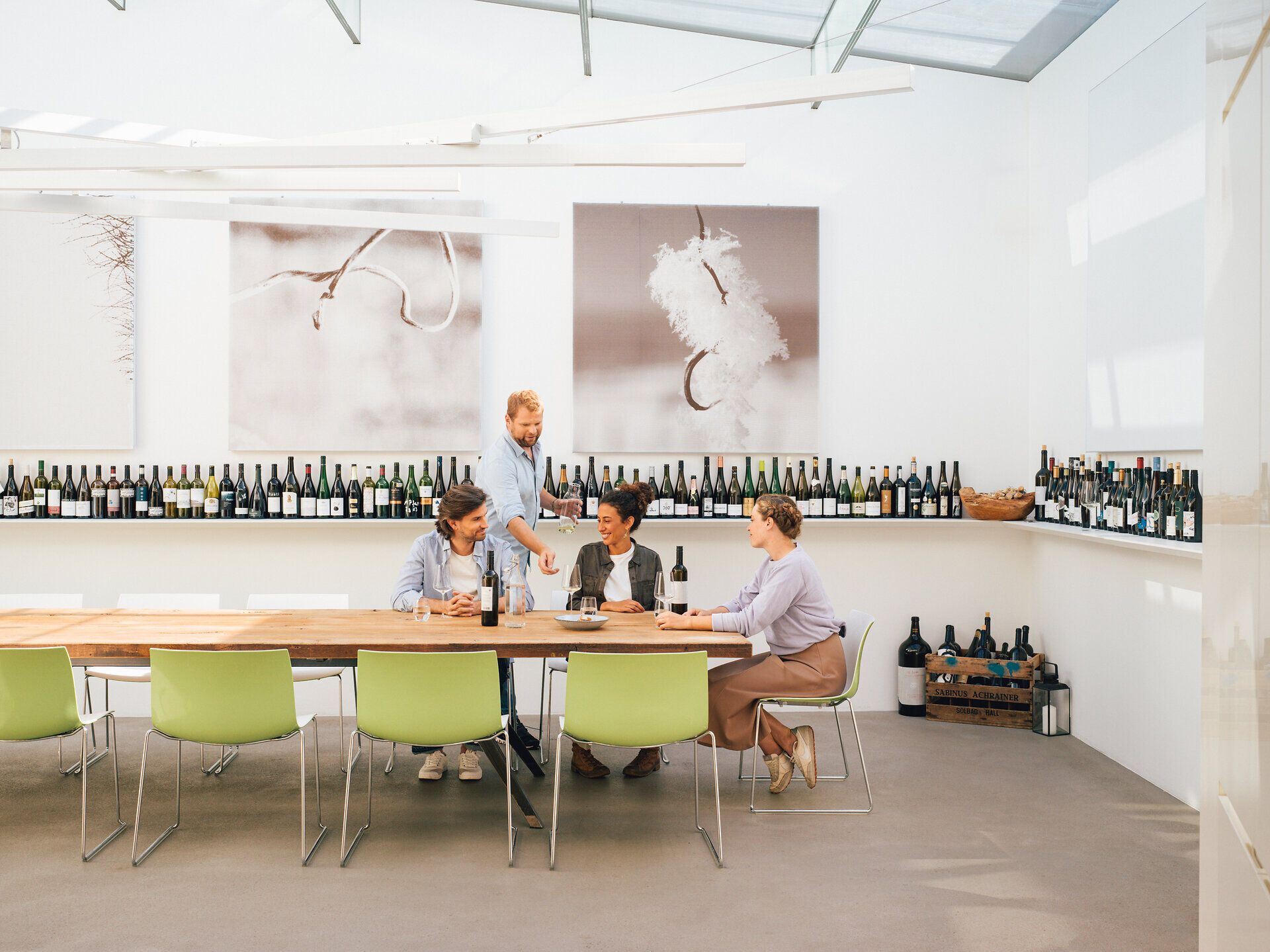 In einem einladenden Raum voller Weinflaschen genießen Freunde die herbstliche Atmosphäre bei einem Glas edlen Weins. Der große Tisch aus Holz lädt zum Verweilen ein und schafft eine gesellige Stimmung, während die sanften Farben der Natur durch die Fenster strömen.