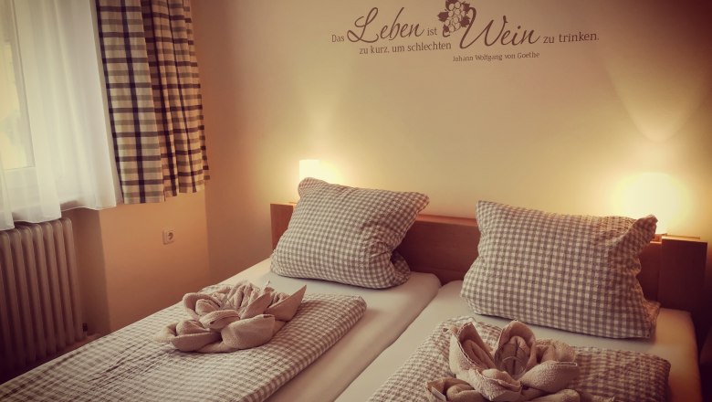 Gemütliches Gästezimmer mit zwei Betten, karierten Bettdecken und Kissen, dekorativen Handtüchern und einem Goethe-Zitat an der Wand.