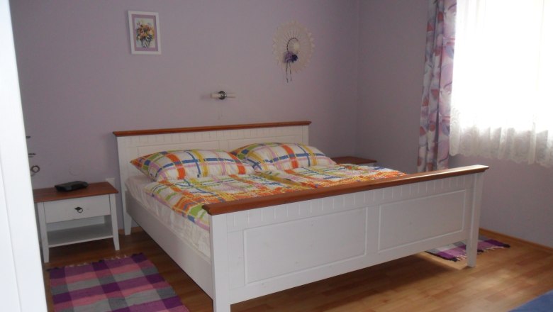 Schlafzimmer, © Fam. Faber Ein Schlafzimmer mit einem weißen Bett, bunten Bettdecken, einem Nachttisch und einem Fenster mit Vorhängen.