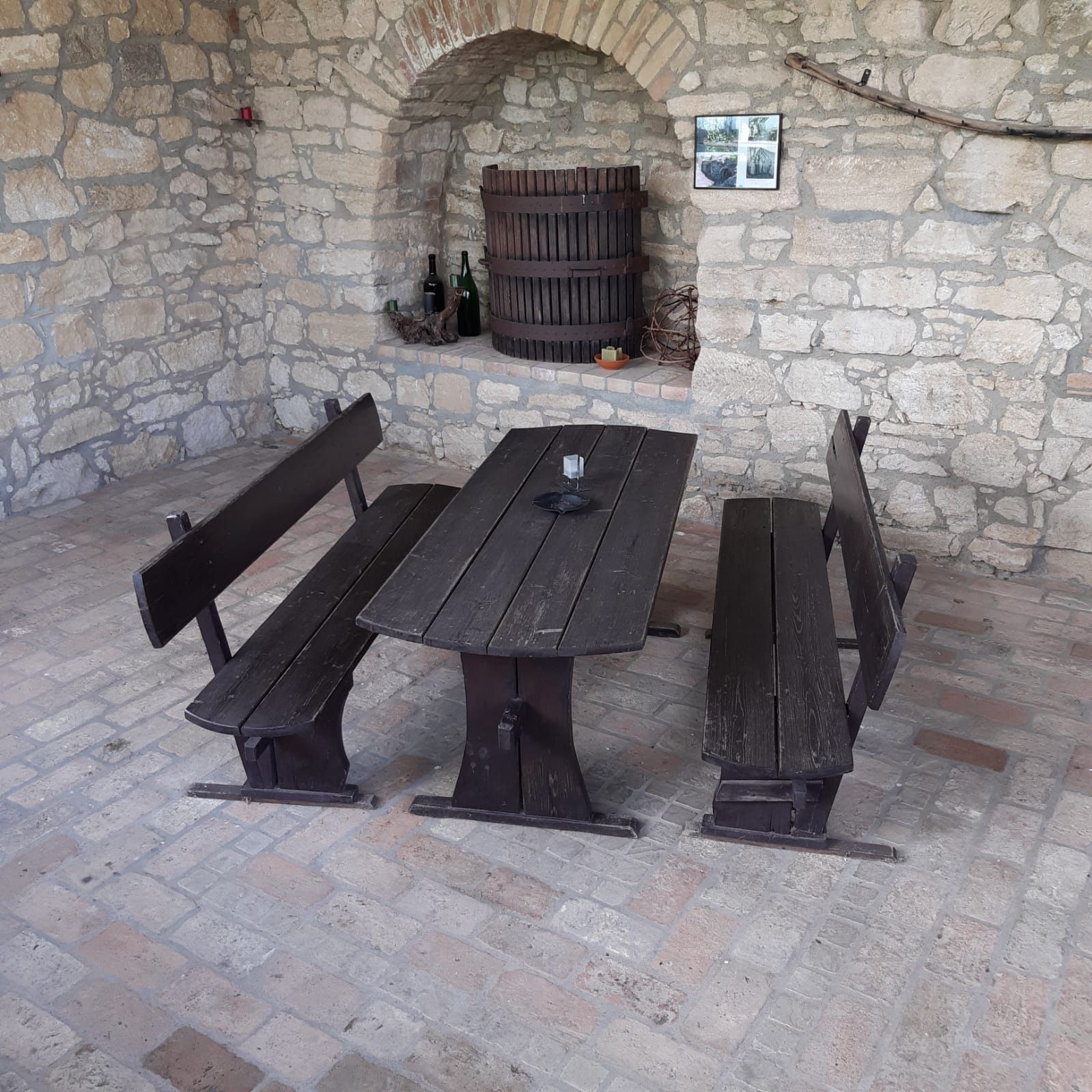 Rustikaler Sitzbereich mit Holztisch und Bänken vor einer Steinwand mit Weinfass.