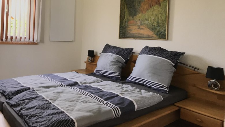 Ein gem&uuml;tliches Schlafzimmer mit einem Doppelbett, grauer Bettw&auml;sche, einem Gem&auml;lde an der Wand und einem Fenster mit Jalousien.