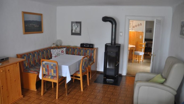 Gem&uuml;tliche Essecke mit Eckbank, Tisch und St&uuml;hlen, daneben ein Ofen. Im Hintergrund ein Durchgang zu einem weiteren Raum.