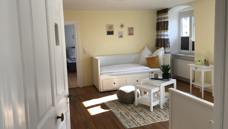 Heller Raum mit gelben W&auml;nden, einem wei&szlig;en Tagesbett, kleinem Tisch und Pflanzen.