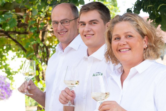 Drei Personen in wei&szlig;er Kleidung halten Weingl&auml;ser und l&auml;cheln in die Kamera.