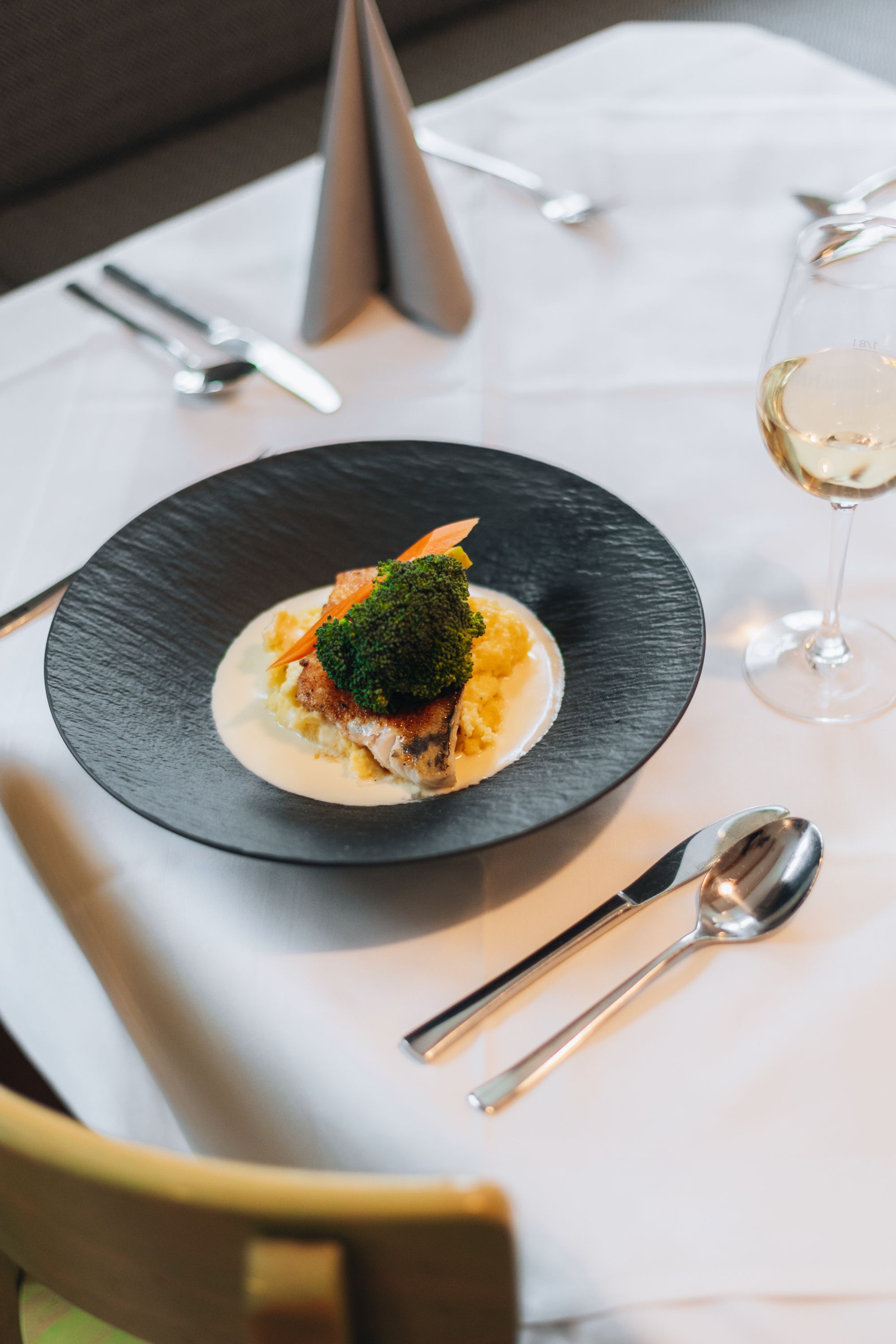 Ein Teller mit einem Gericht aus Fisch, Brokkoli und Kartoffelpüree auf einem gedeckten Tisch mit Besteck und einem Glas Weißwein.