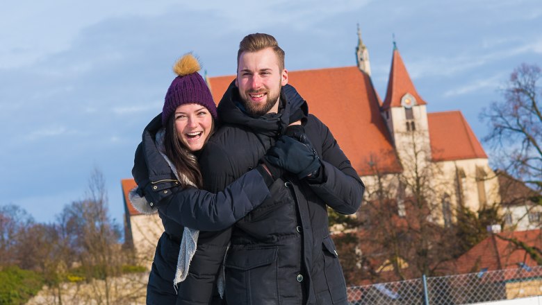 A couple on an ice rink, in the background a church with a red roof.