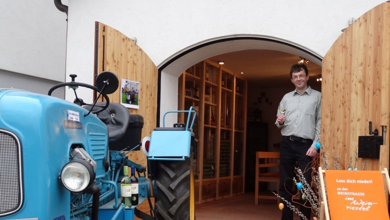 Ein Mann steht mit einem Weinglas in der Hand vor einem Gebäude mit offenen Holztüren. Ein blauer Traktor steht daneben.