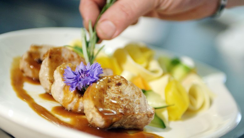 Ein Teller mit Fleisch, Soße, Nudeln und Gemüse, dekoriert mit einer Blume und Rosmarin.