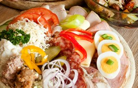 Verschiedene Wurst- und Käsesorten auf einem Holzbrett, garniert mit Gemüse und Eiern, daneben ein Salat in einer Glasschüssel.