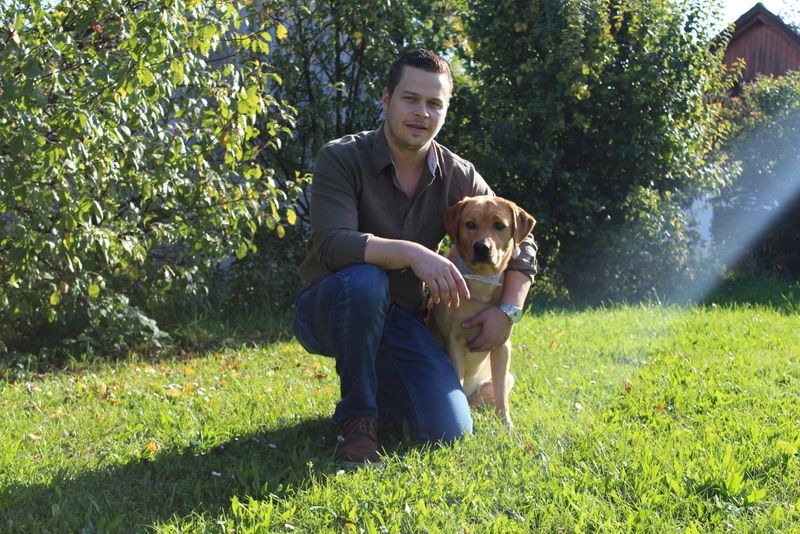 Ein Mann kniet auf einer Wiese neben einem Hund, umgeben von Bäumen und Sonnenlicht.
