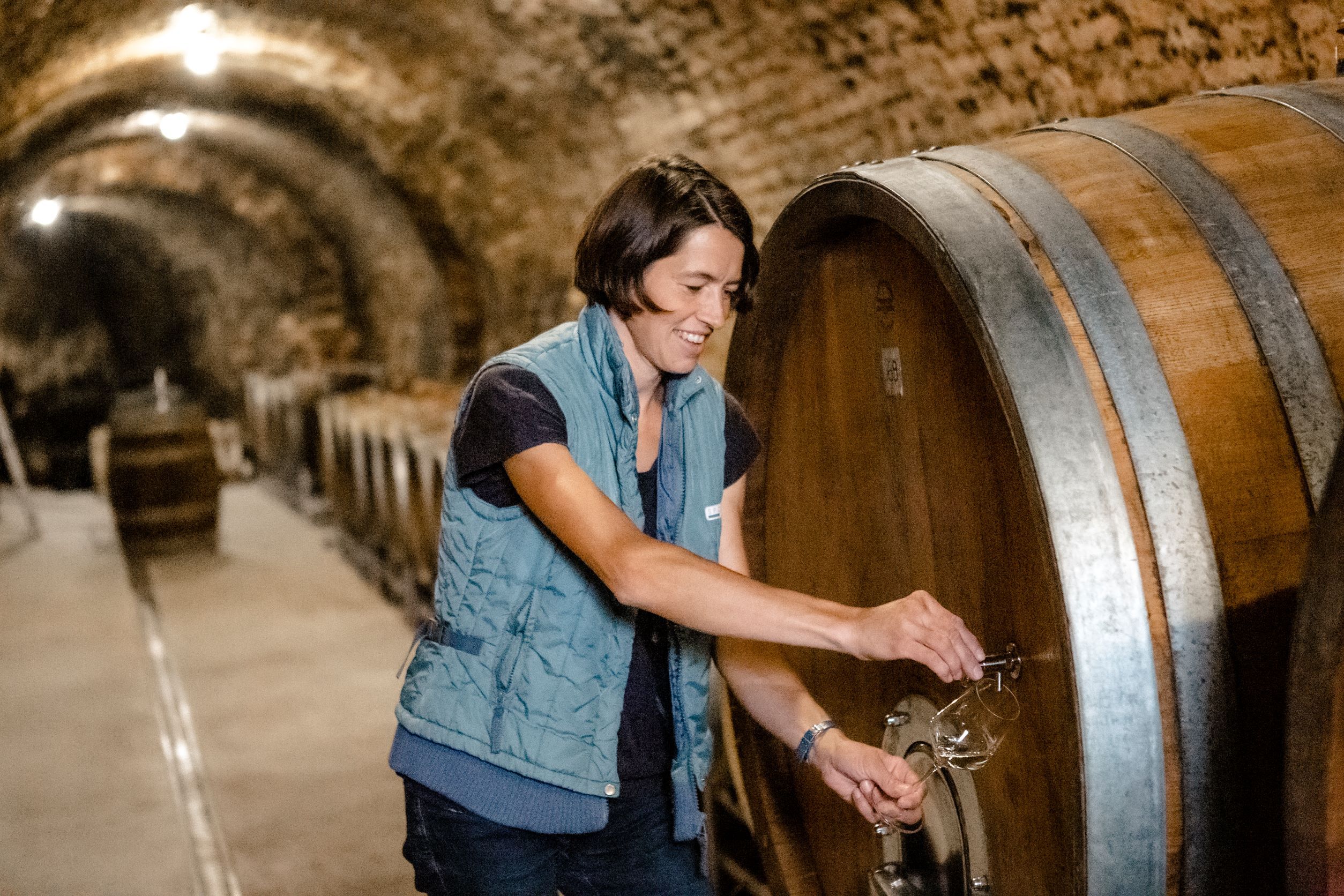 A person in a winery draws wine from a large wooden barrel.