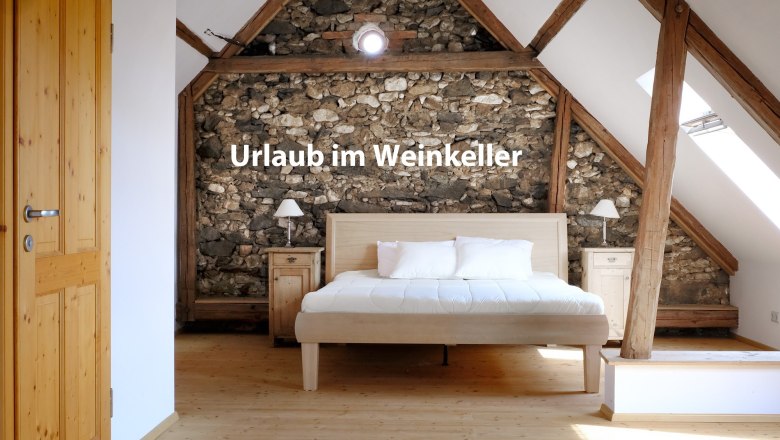 Ferienhaus "Weinkeller Röschitz", © Weiss und Waldschmidt Schlafzimmer mit Holzbalken und Steinwand, Bett in der Mitte, Text 'Urlaub im Weinkeller'.