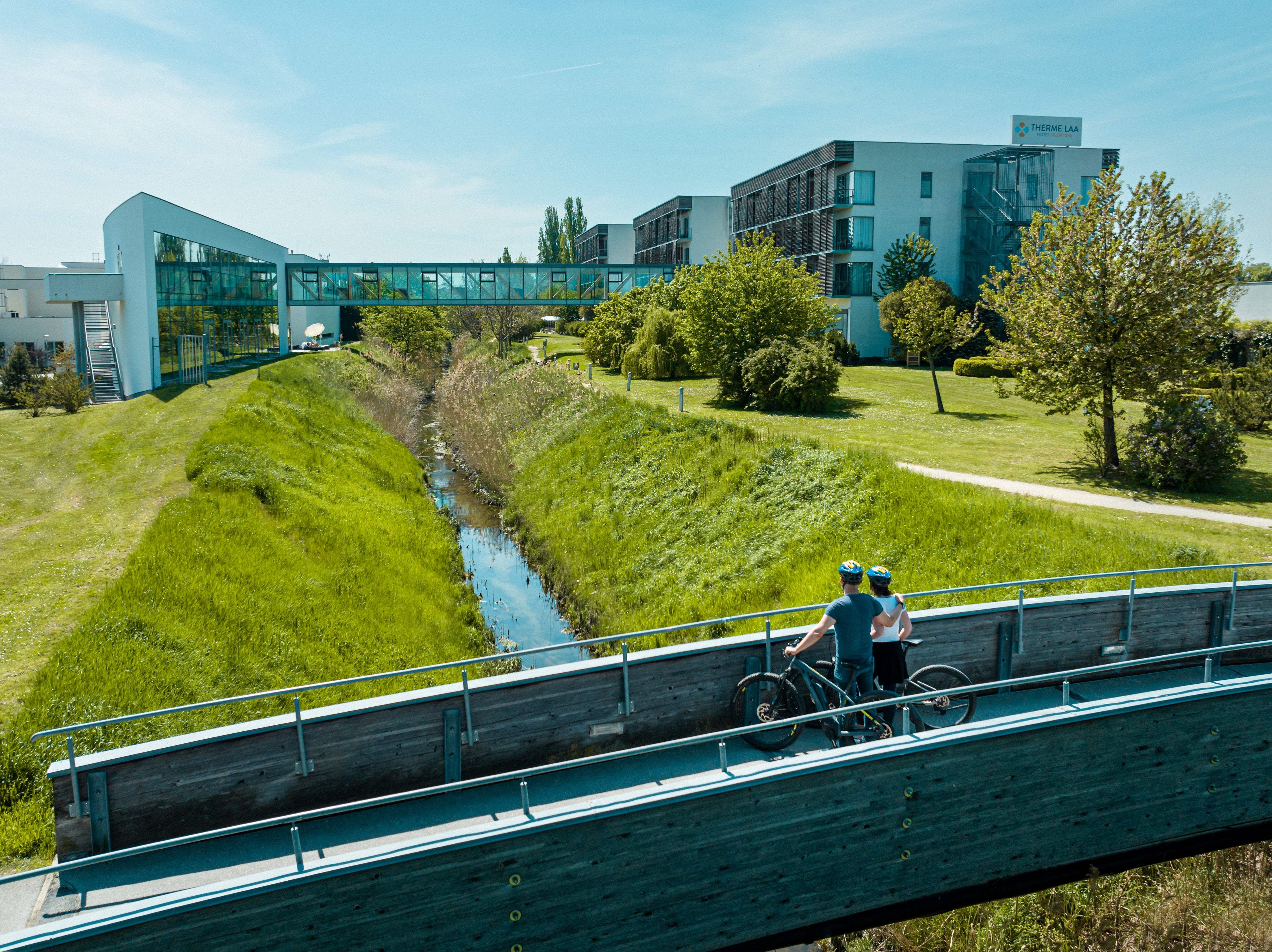 Zwei Radfahrer auf einer Brücke vor der Therme Laa & Hotel.
