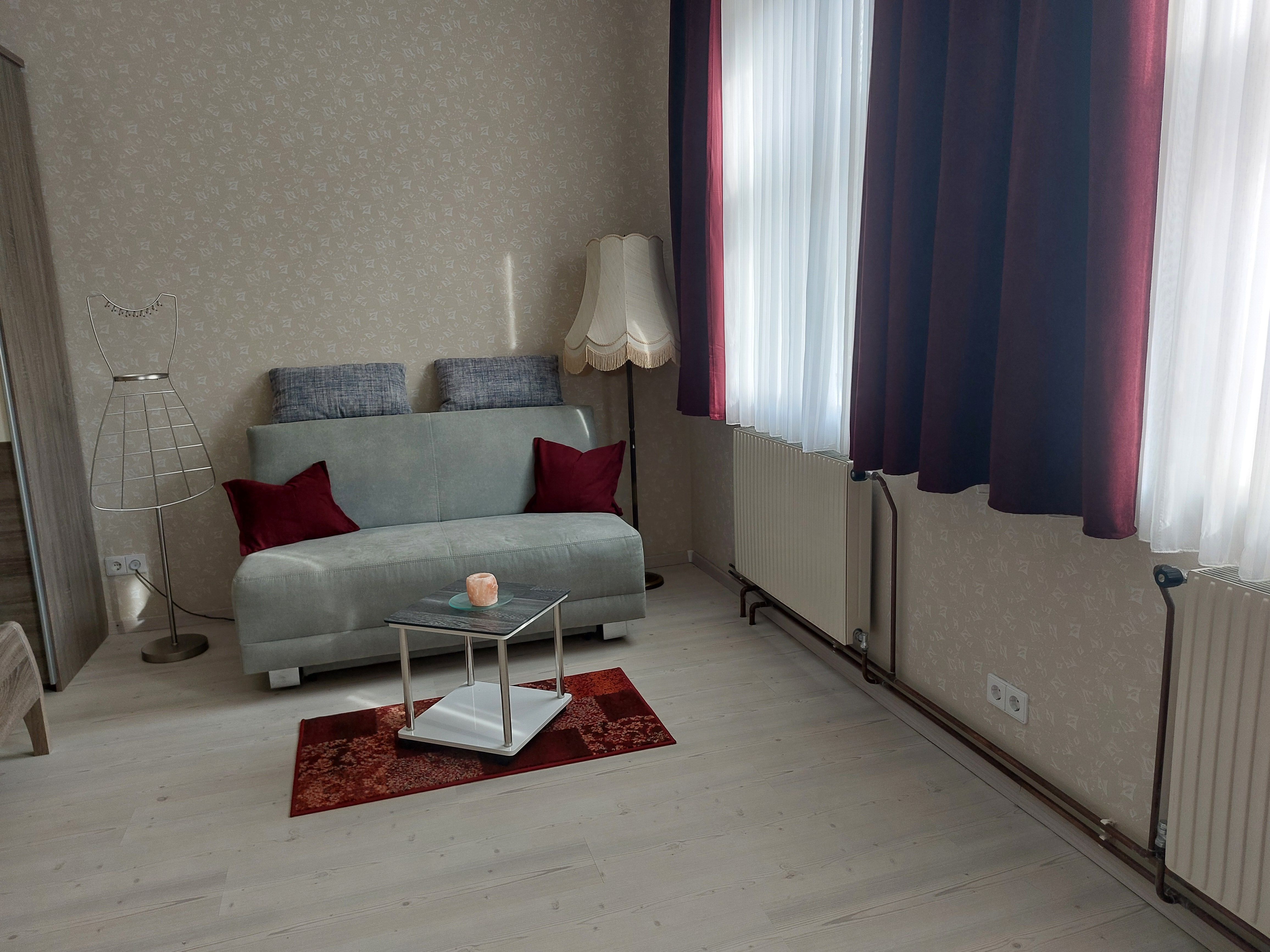 Wohnzimmer mit grauem Sofa, roten Kissen, Stehlampe und kleinem Tisch auf rotem Teppich.