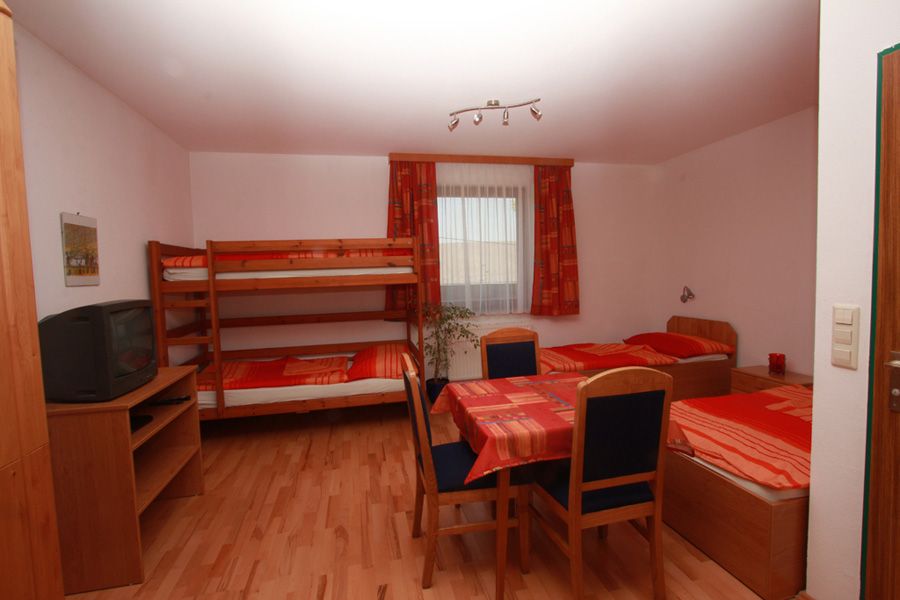 Ein einfach eingerichtetes Zimmer mit Etagenbett, Einzelbett, Tisch und Stühlen. Ein Fernseher steht auf einem Regal. Fenster mit Vorhängen im Hintergrund.