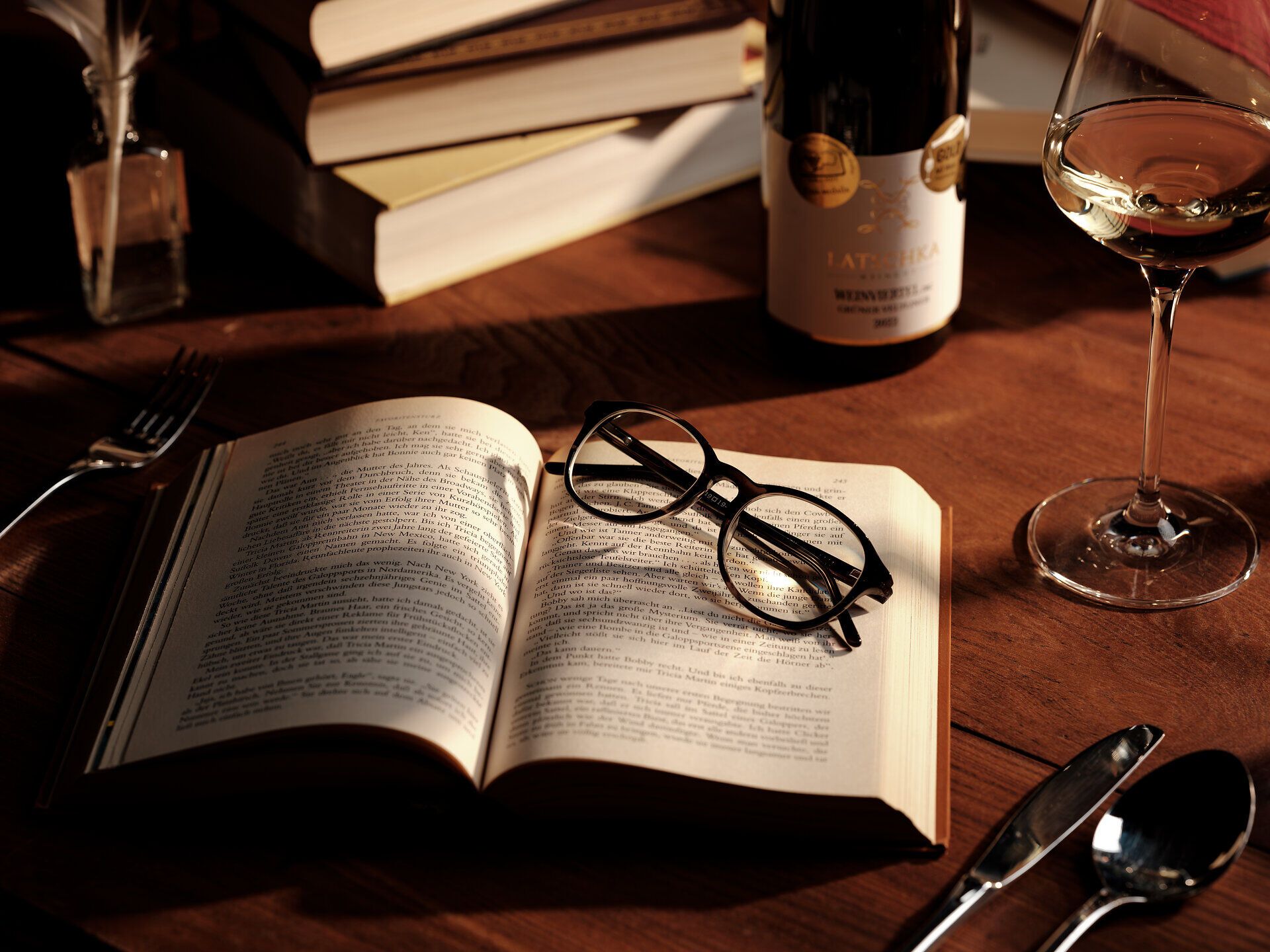 Einladend liegt ein aufgeschlagenes Buch auf einem rustikalen Holztisch, umgeben von einer Flasche Wein und einem Glas. Die sanften Lichtstrahlen schaffen eine gemütliche Atmosphäre, die zum Verweilen und Genießen einlädt. Hier vereinen sich Kulinarik und Kultur in harmonischer Stille.