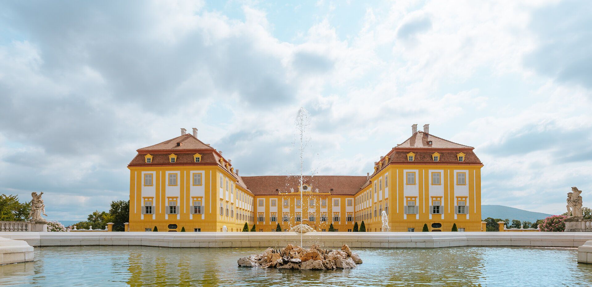 Die majestätische Fassade des Schlosses Hof strahlt in warmen Gelbtönen und spiegelt sich im ruhigen Wasser des Brunnens wider. Umgeben von üppigen Gärten und Terrassen lädt dieser Ort dazu ein, die Schönheit der Natur und die Pracht der Architektur zu genießen.