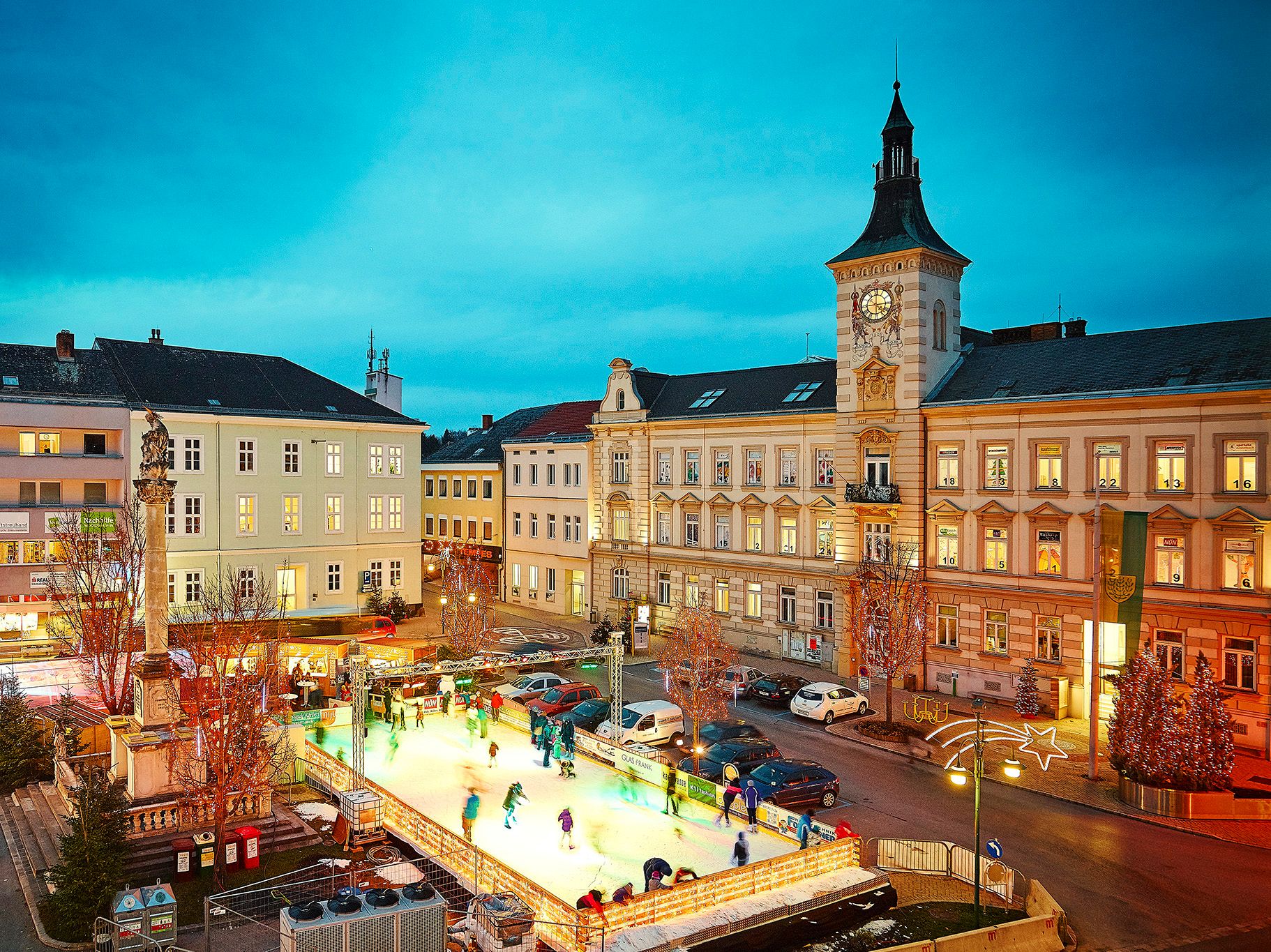 Eislaufplatz in einer Stadt bei Abenddämmerung, umgeben von historischen Gebäuden und Weihnachtsbeleuchtung.