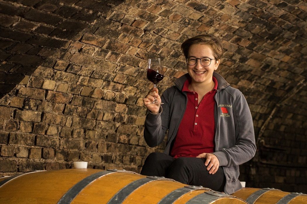 Eine Winzerin steht mit einem Glas Rotwein in der Hand im Weinkeller umgeben von Holzfässern.