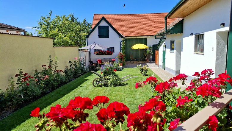 Ein gepflegter Garten mit roten Blumen, einem alten Pflug und einem Haus mit roten Dachziegeln im Hintergrund.