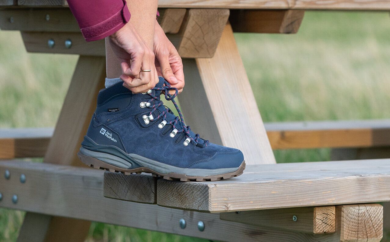 Eine Person hat den Fuß auf einer Holzbank abgestellt und schnürt den Wanderschuh zu.