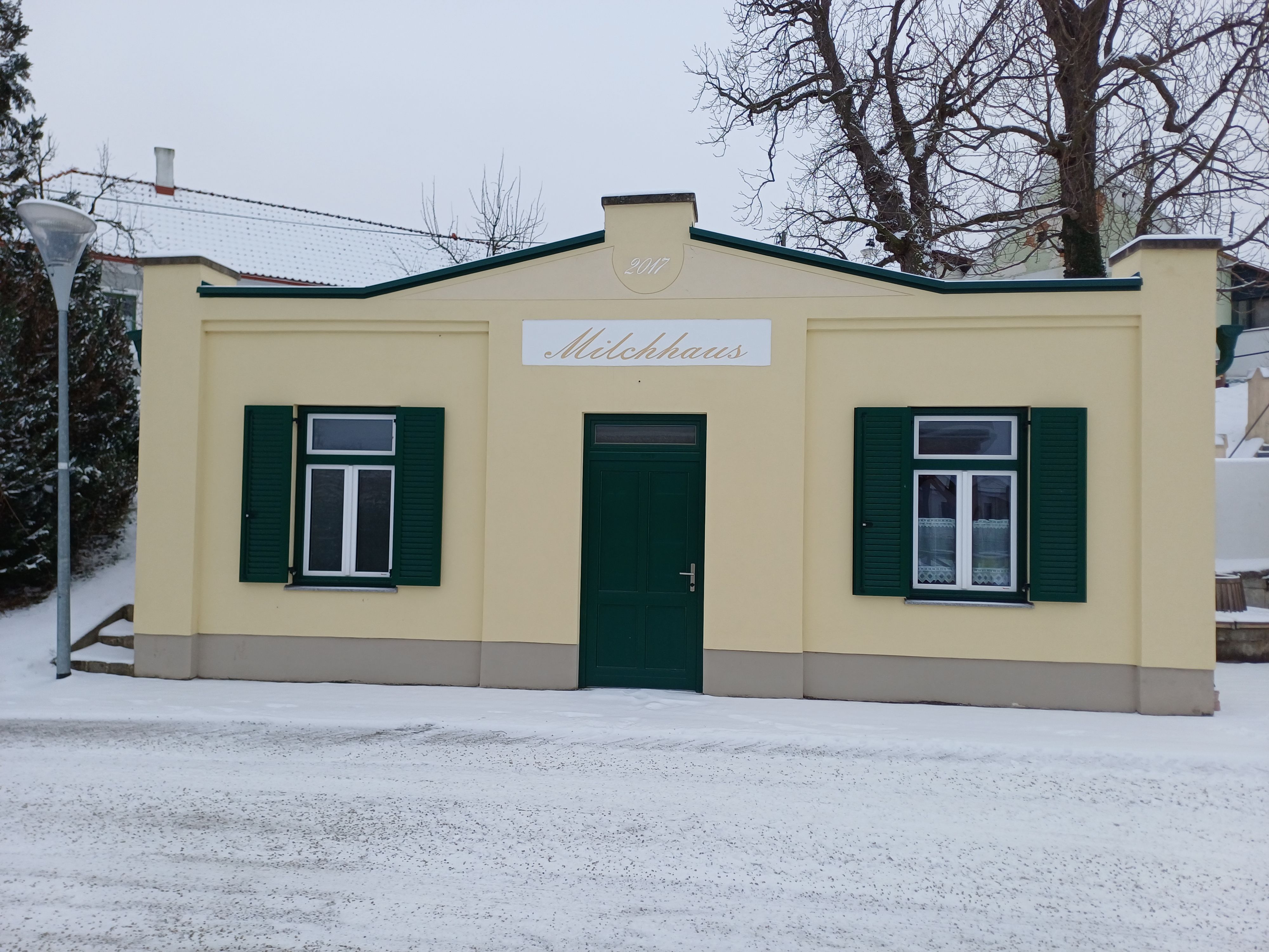 Malá budova s nápisem "Milchhaus" se zelenými okenicemi, obklopená sněhem.