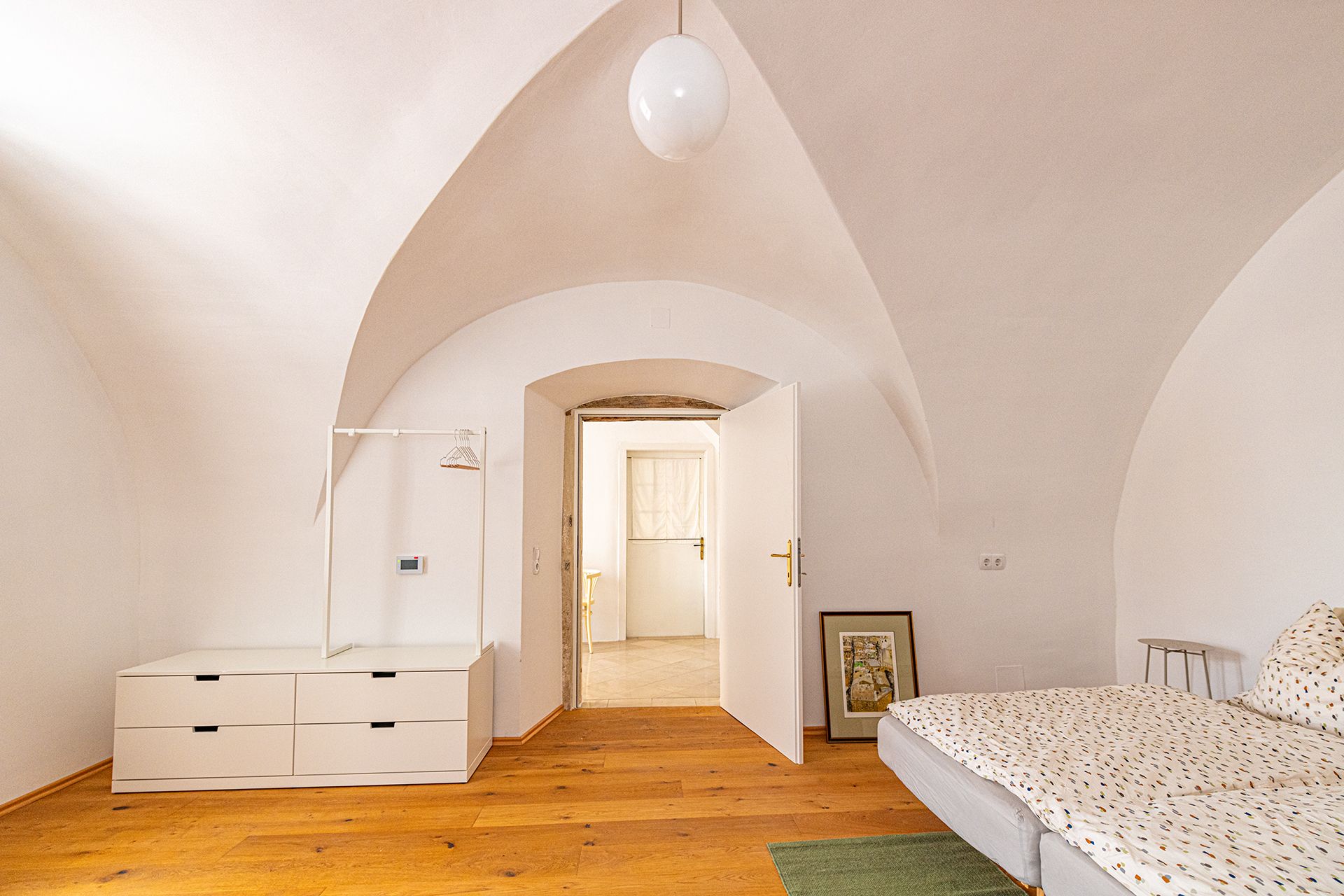Ein Zimmer mit Gewölbedecke, Holzboden, Bett, Kommode und offener Tür.