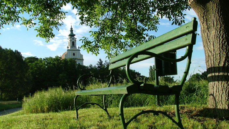 Eine grüne Bank unter einem Baum mit Blick auf eine Kirche im Hintergrund.