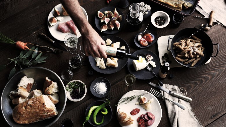 A rustic table with various dishes, including bread, cheese, figs, sausage and fish. A hand pours oil over a dish.