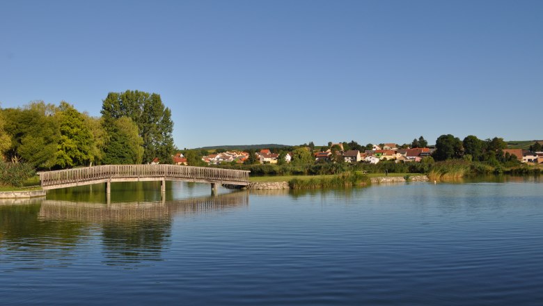 Holzbrücke über einen Teich mit Dorf im Hintergrund.