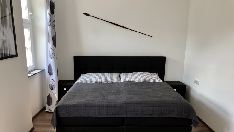 Modernes Schlafzimmer mit einem gro&szlig;en Bett, grauer Bettdecke und einem Speer an der Wand.