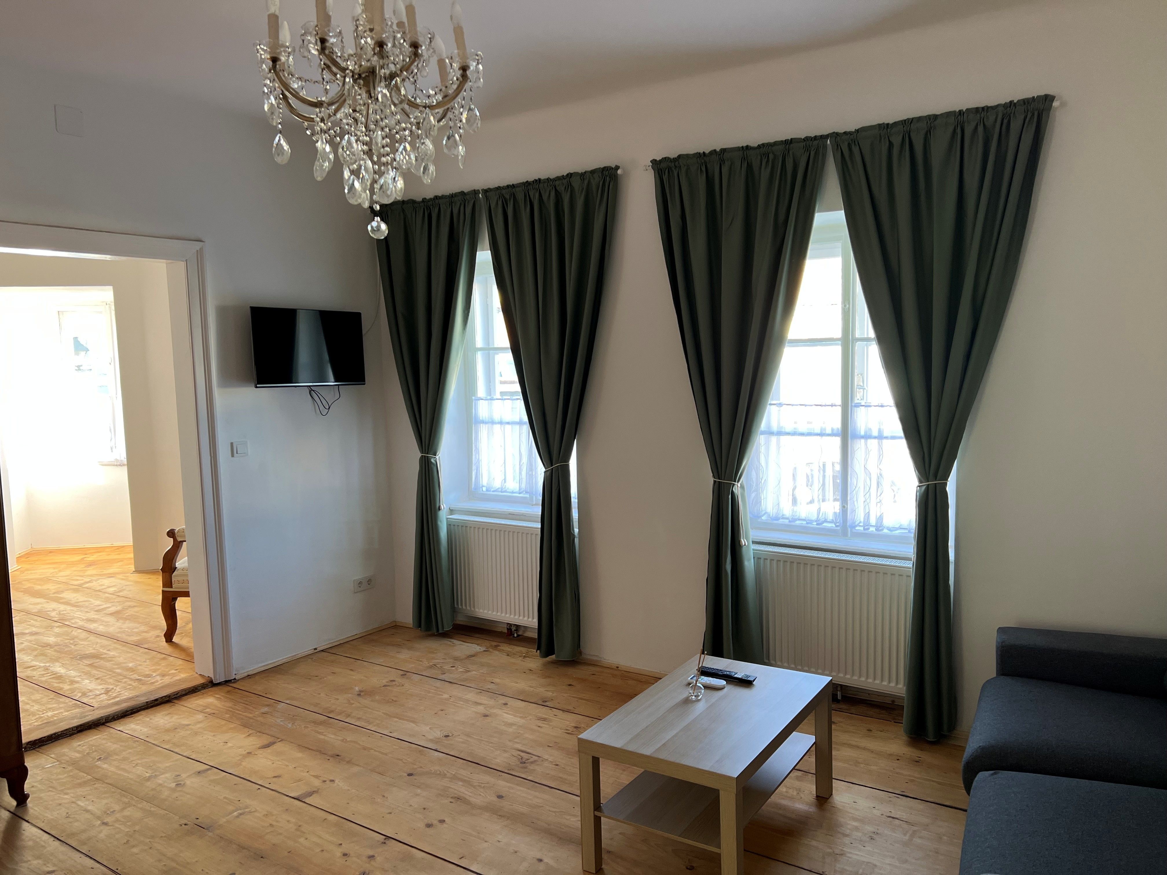 Ein Wohnzimmer mit Holzboden, einem Kronleuchter, zwei Fenstern mit grünen Vorhängen, einem grauen Sofa und einem kleinen Holztisch.