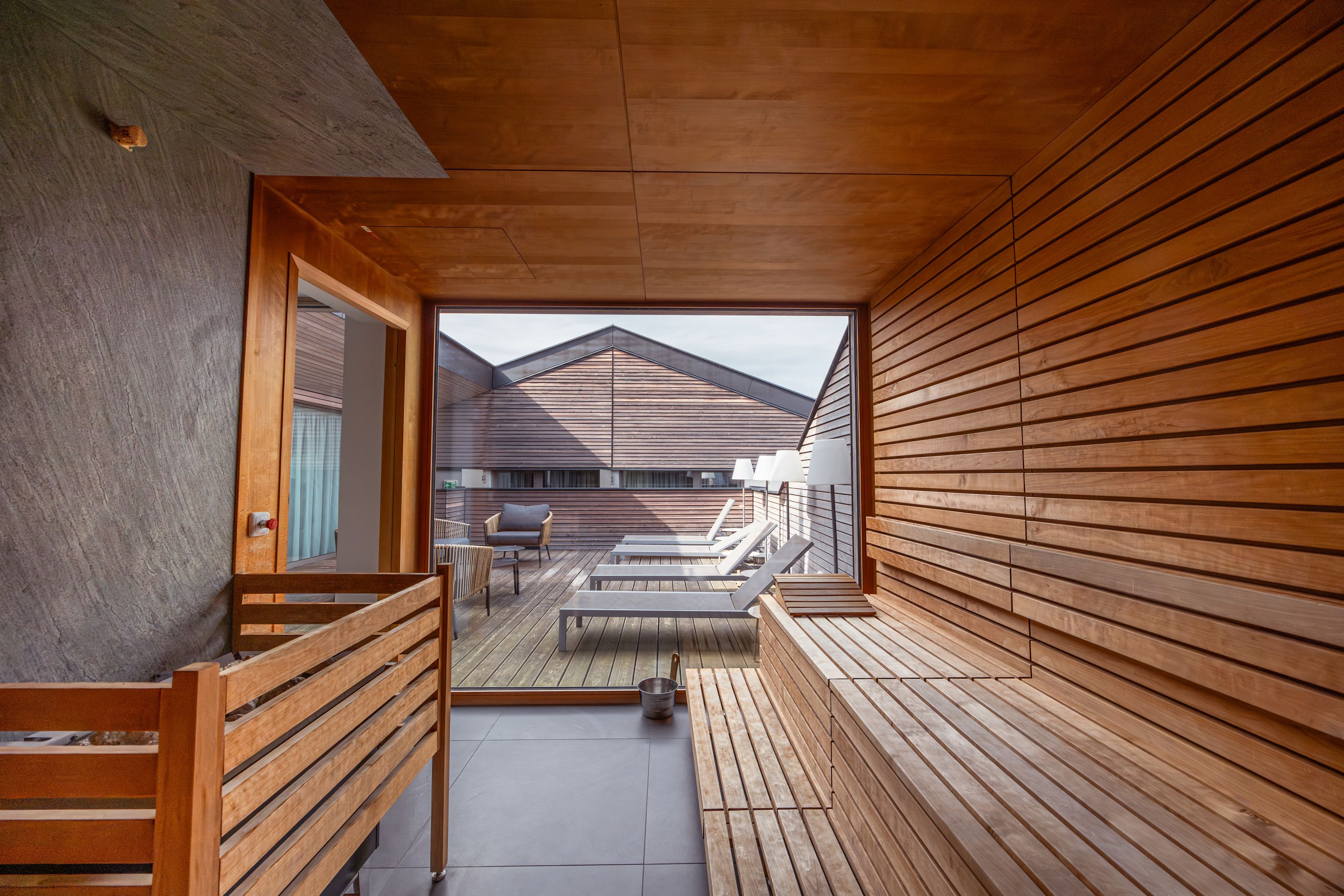Innenansicht einer Sauna mit Blick auf eine Terrasse mit Liegestühlen und Holzwänden.
