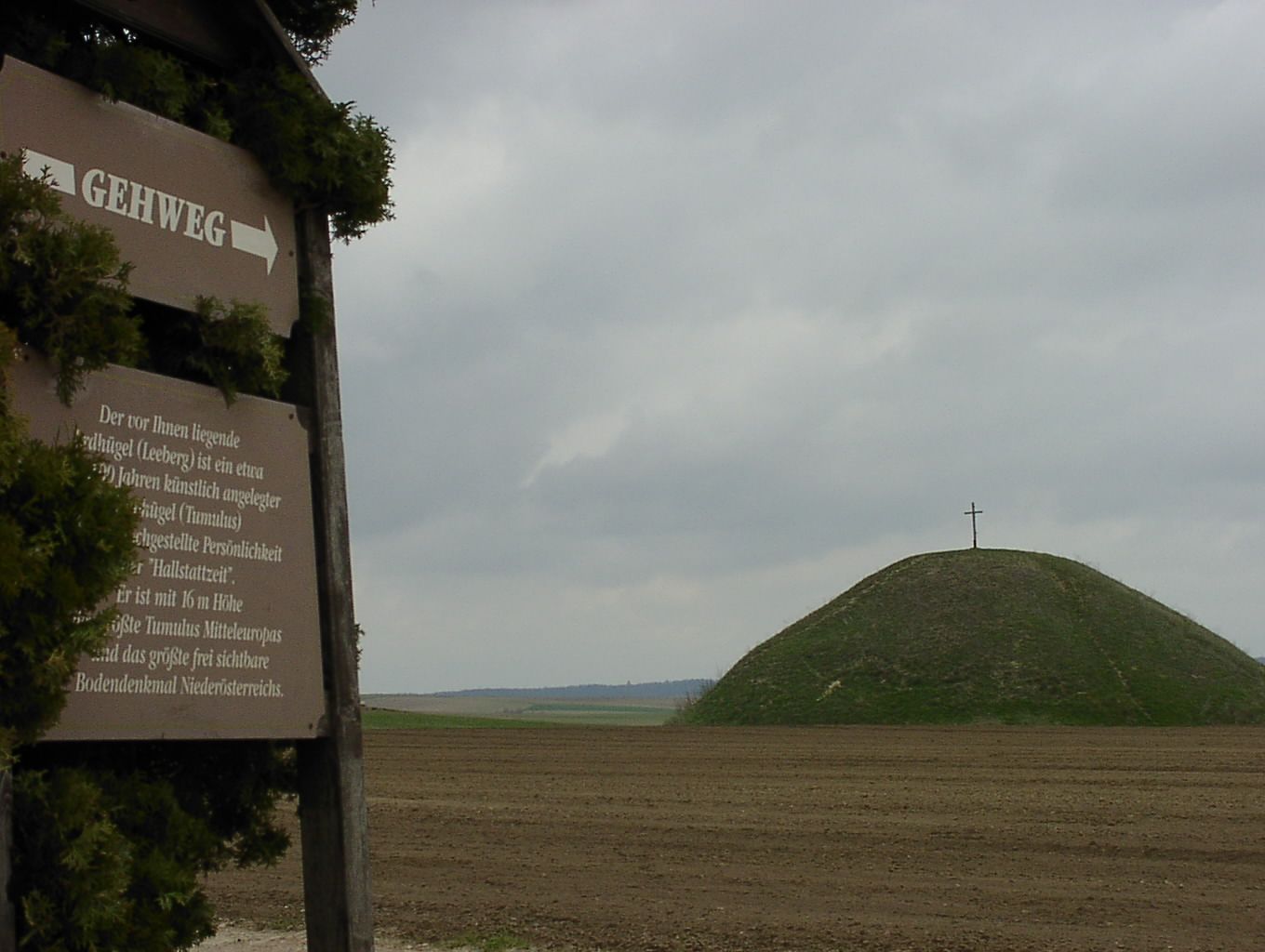 Ein Hügel mit einem Kreuz auf der Spitze, daneben ein Schild mit der Aufschrift 'Gehweg'.