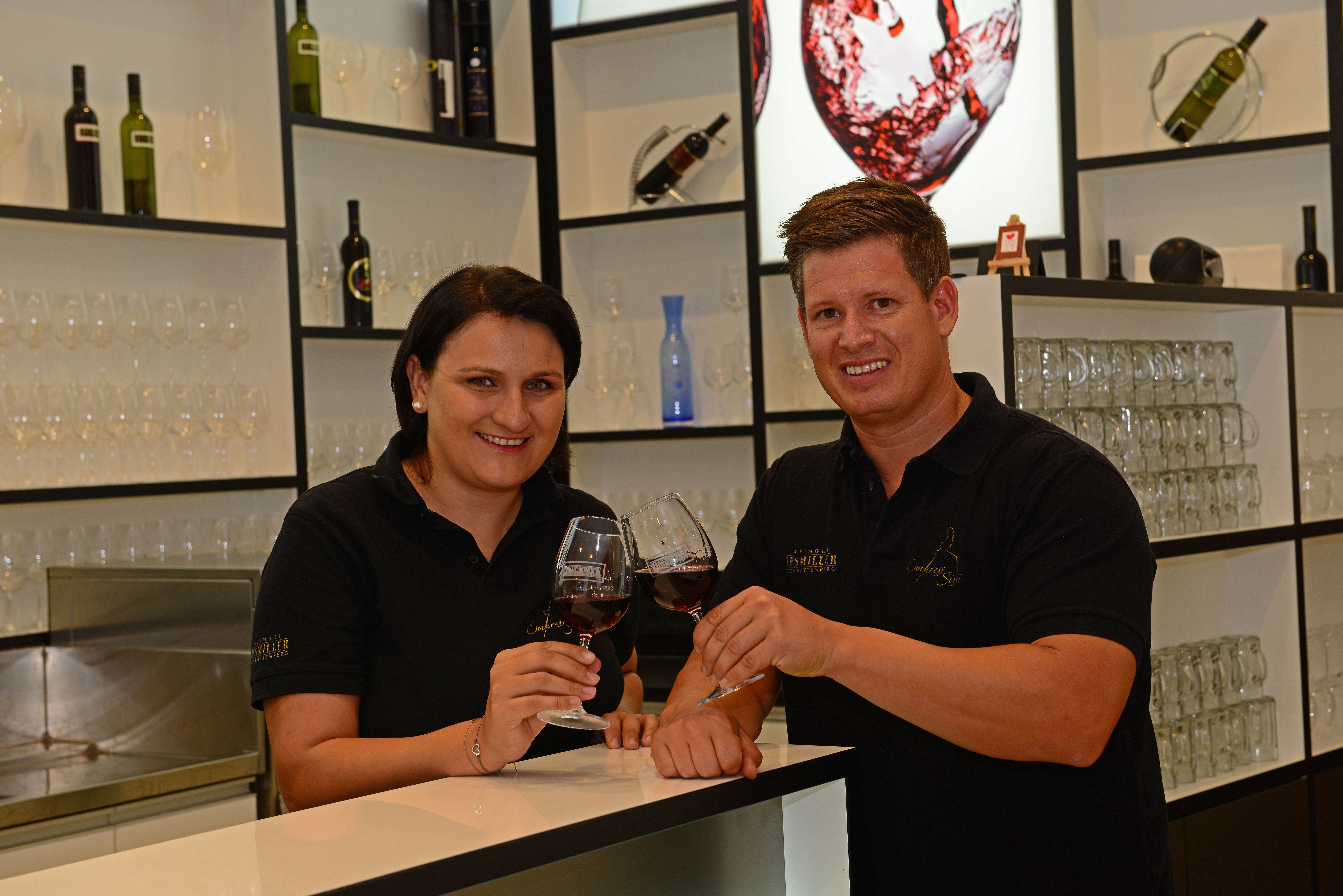 Ein Paar steht in einem Verkostungsraum, es wird mit einem Glas Rotwein angestoßen.