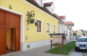 Gelbes Gebäude mit Schild 'Gästezimmer Buschenschank Weinbau Poys' und geparktem Auto davor.