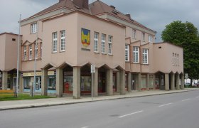 Ein modernes Rathausgeb&auml;ude mit rosa Fassade und gro&szlig;en Fenstern. Es hat ein Wappen an der Ecke und Gesch&auml;fte im Erdgeschoss.