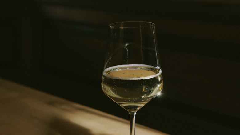 A wine glass with white wine stands on a wooden table in the shade.