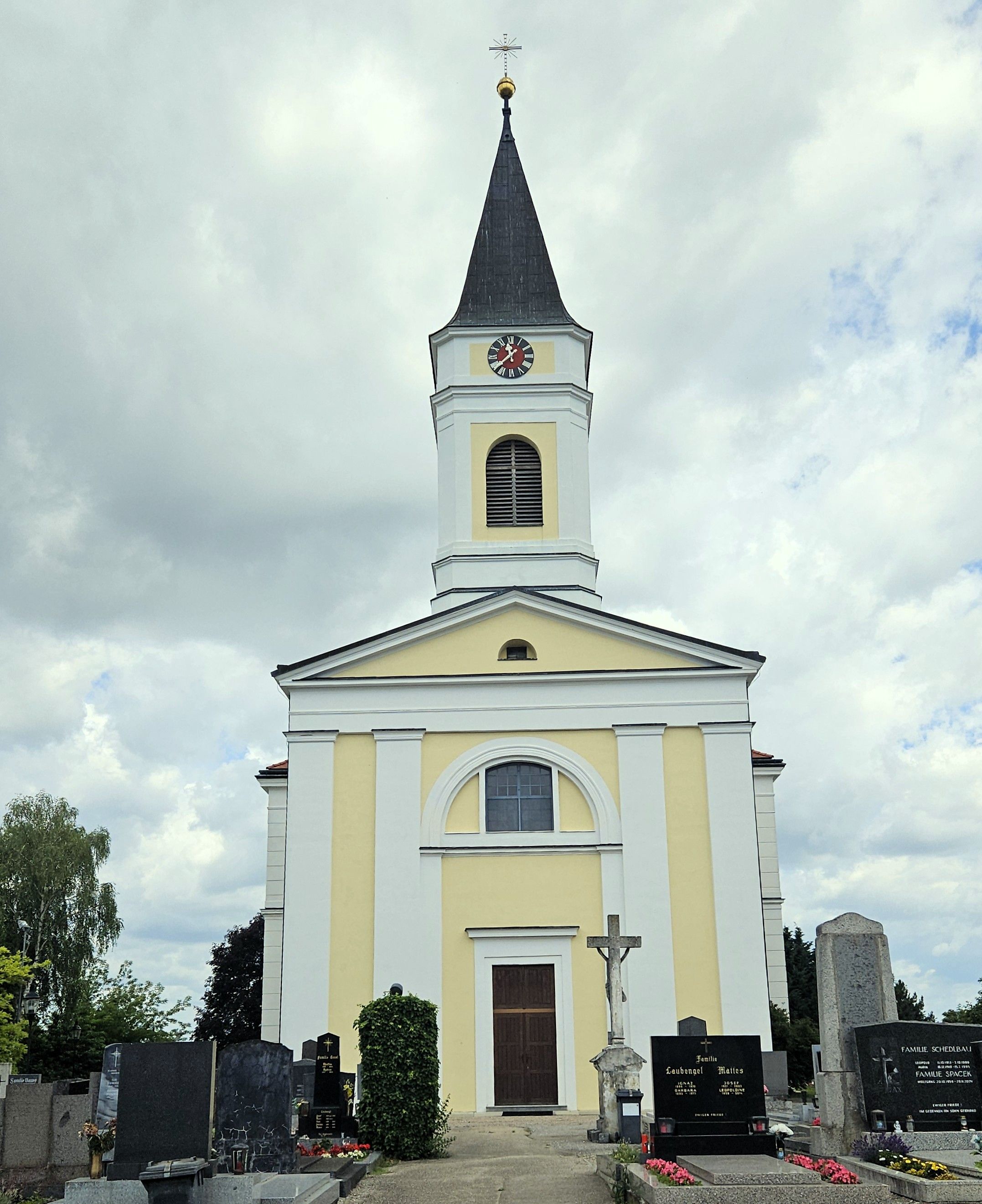 Pfarrkirche in Kammersdorf mit hohem Turm, Kreuz und Friedhof im Vordergrund unter bewölktem Himmel.