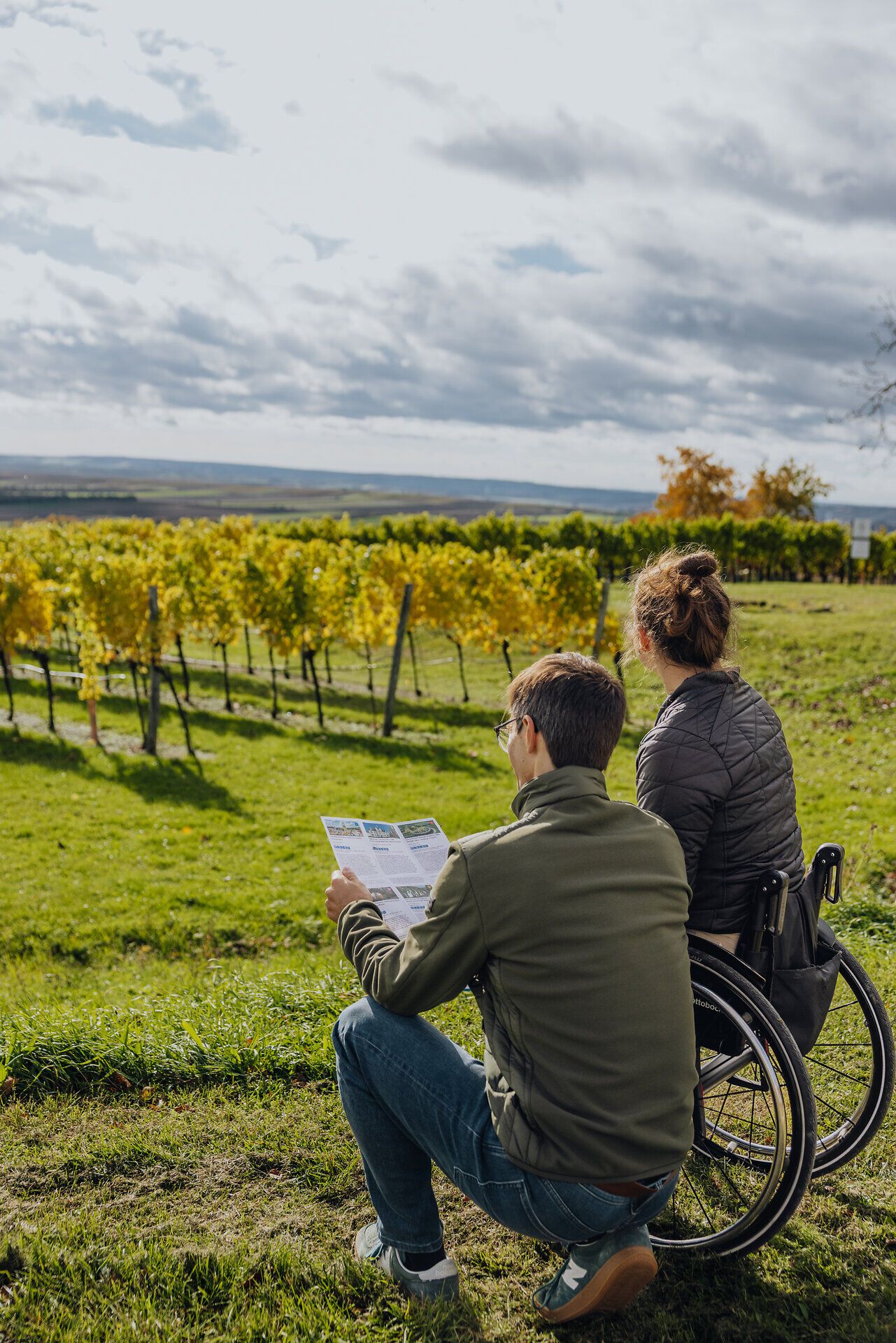 Eine junge Frau im Rollstuhl und ein Mann genießen den Ausblick auf eine mit herbstlich gefärbten Weingärten durchzogene Landschaft. Der Mann hält einen Folder mit Tipps in der Hand.