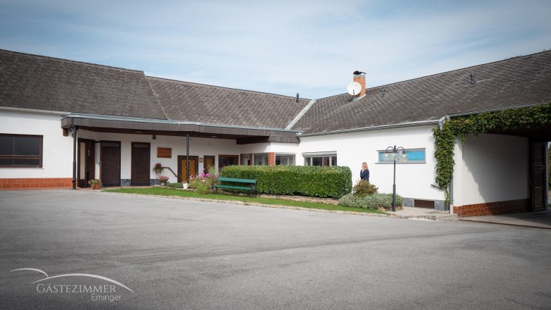 Ein Hof mit einem Gebäude, Parkplätzen und einer Hecke. Eine Person steht neben der Hecke. Auf dem Gebäude ist eine Satellitenschüssel zu sehen.