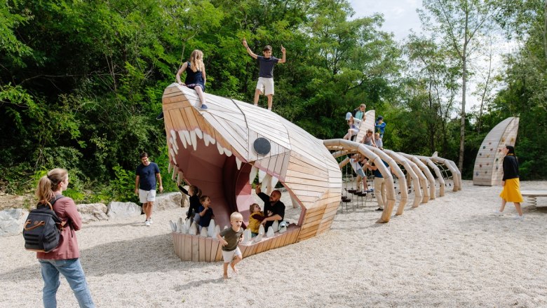 Children play on a large, wooden dinosaur skeleton in a playground.