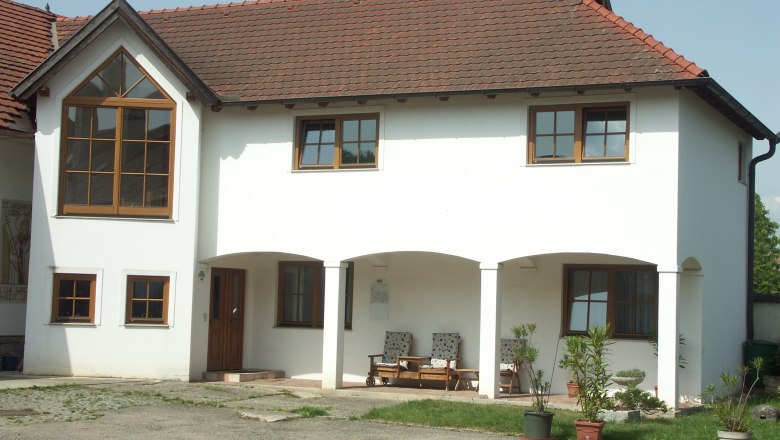 Ein zweistöckiges weißes Gebäude mit roten Dachziegeln und großen Fenstern. Vor dem Haus stehen Pflanzen in Töpfen.