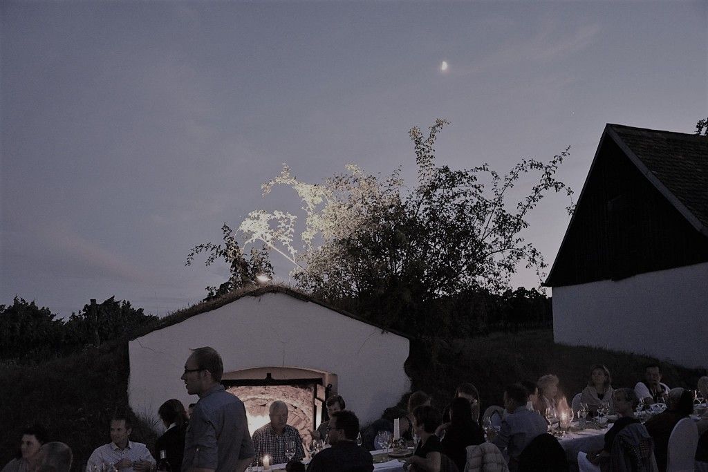 Menschen sitzen an einem langen Tisch im Freien bei Dämmerung, umgeben von Gebäuden und Bäumen.