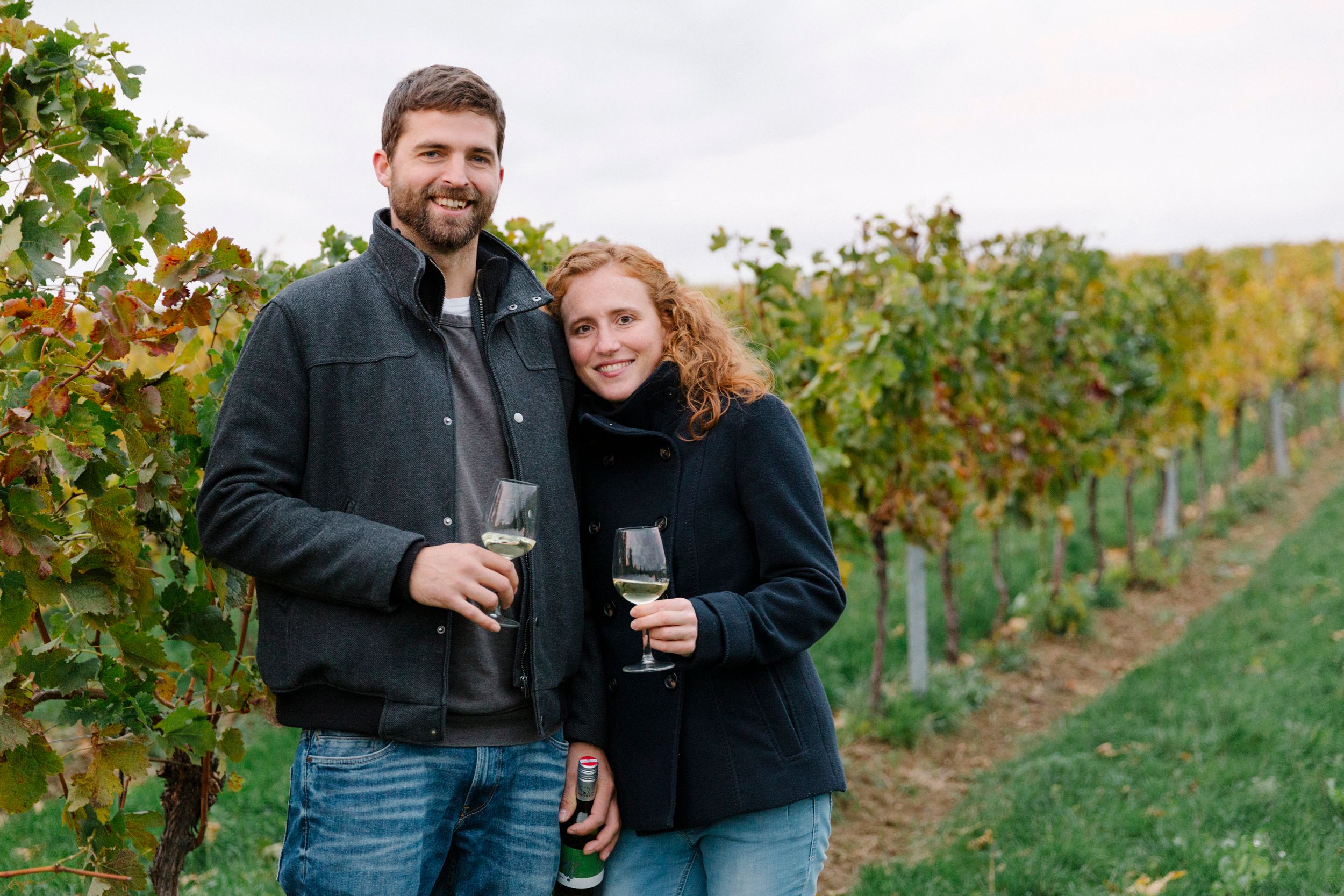 A couple stands in a vineyard holding glasses of wine.